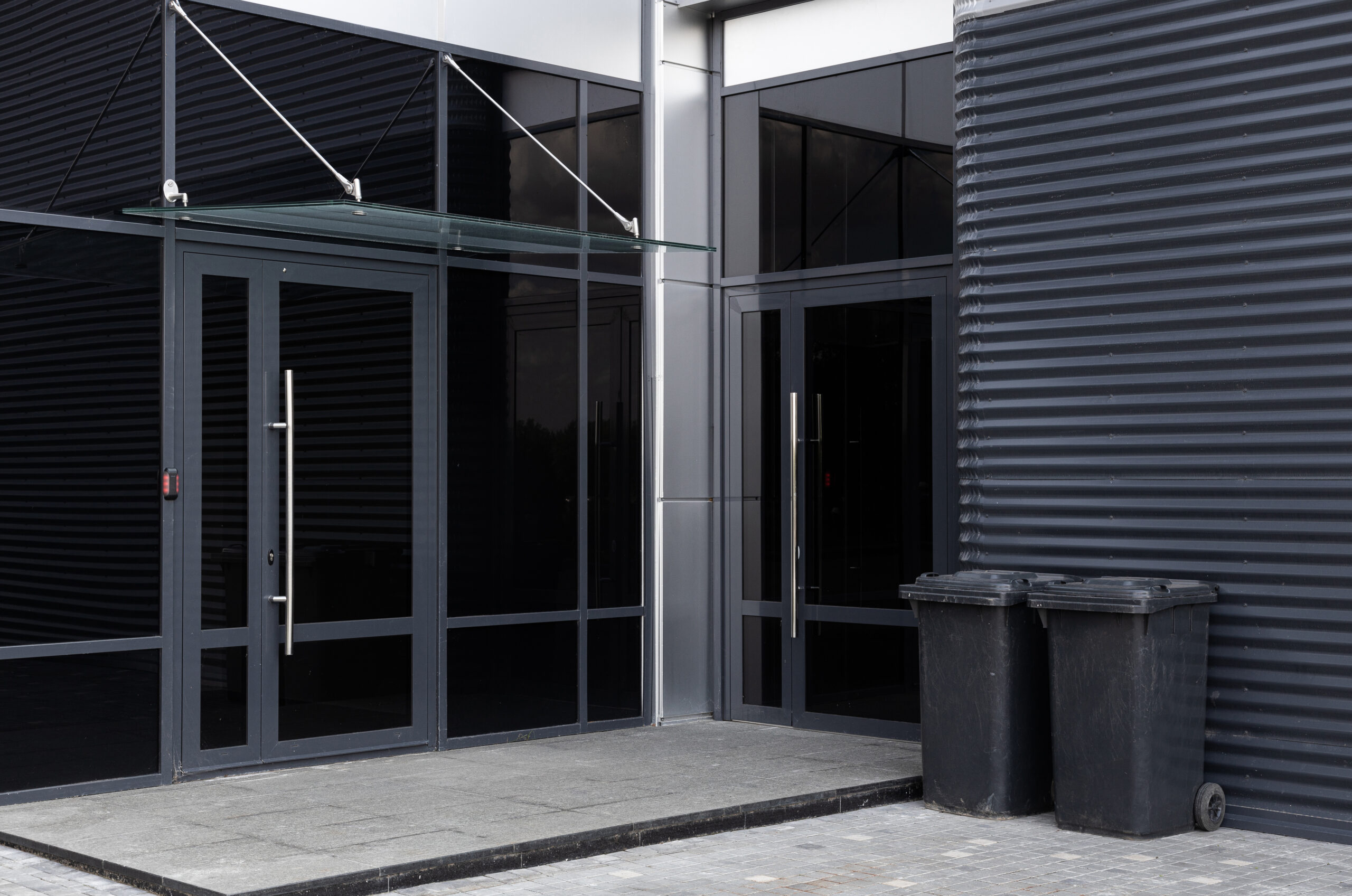 Glass doors entrance to modern building and two scratched black dustbins against black metal corrugated wall nearby